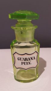 Szklane naczynie aptekarskie (sztanda) barwy uranowej zawierające sproszkowaną guaranę. Roślina ta zawiera 4 razy więcej kofeiny niż ziarna kawy. Indianie z Amazonii nazywają ją „eliksirem młodości”.