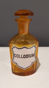 Szklane naczynie aptekarskie (sztanda) barwy oranżowej zawierające kolodium, czyli środek na skaleczenia lub otarcia skóry.